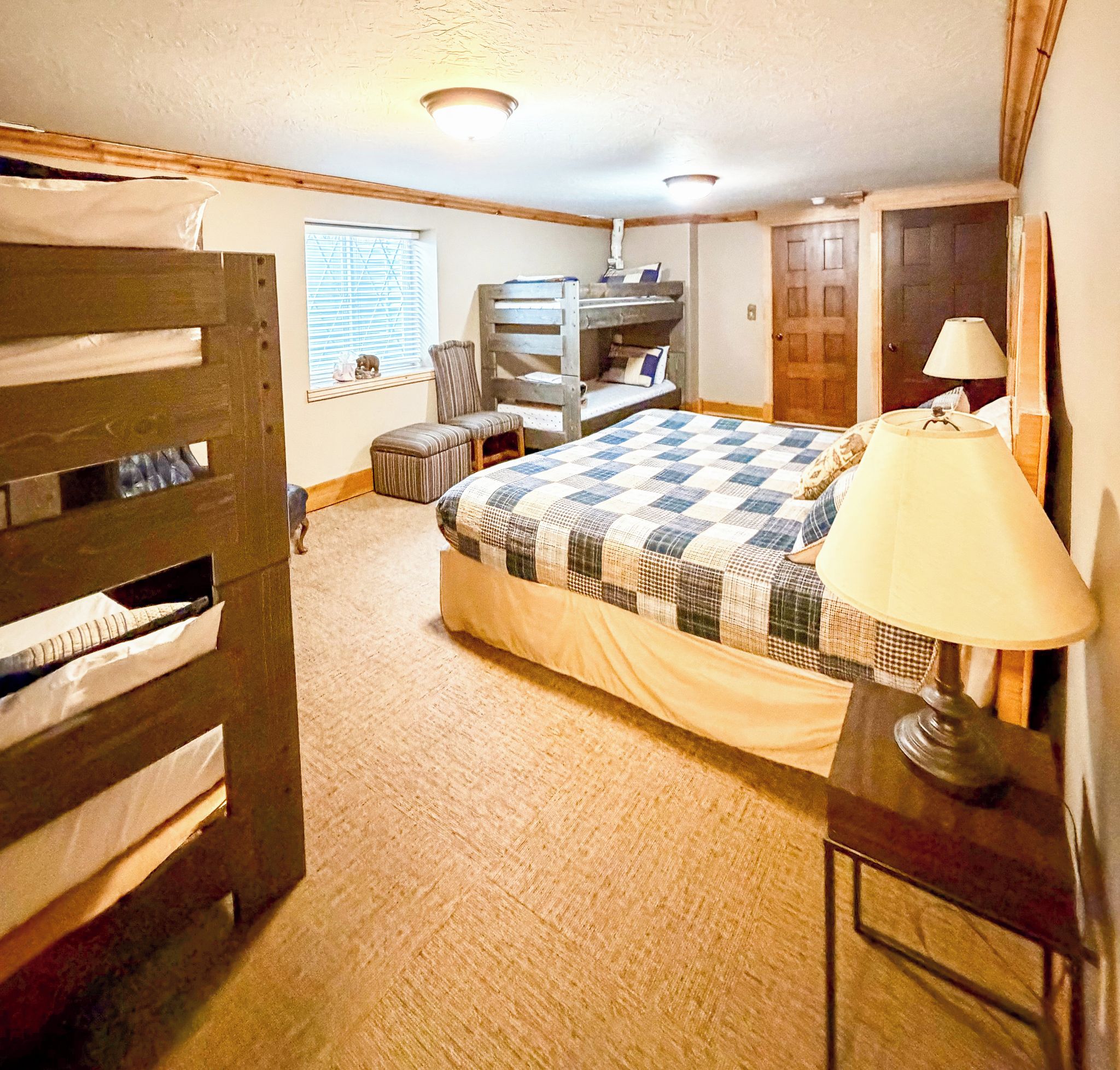 A bedroom with a bunk bed, a chair, a bedside table with a lamp, a window with blinds, and a door.