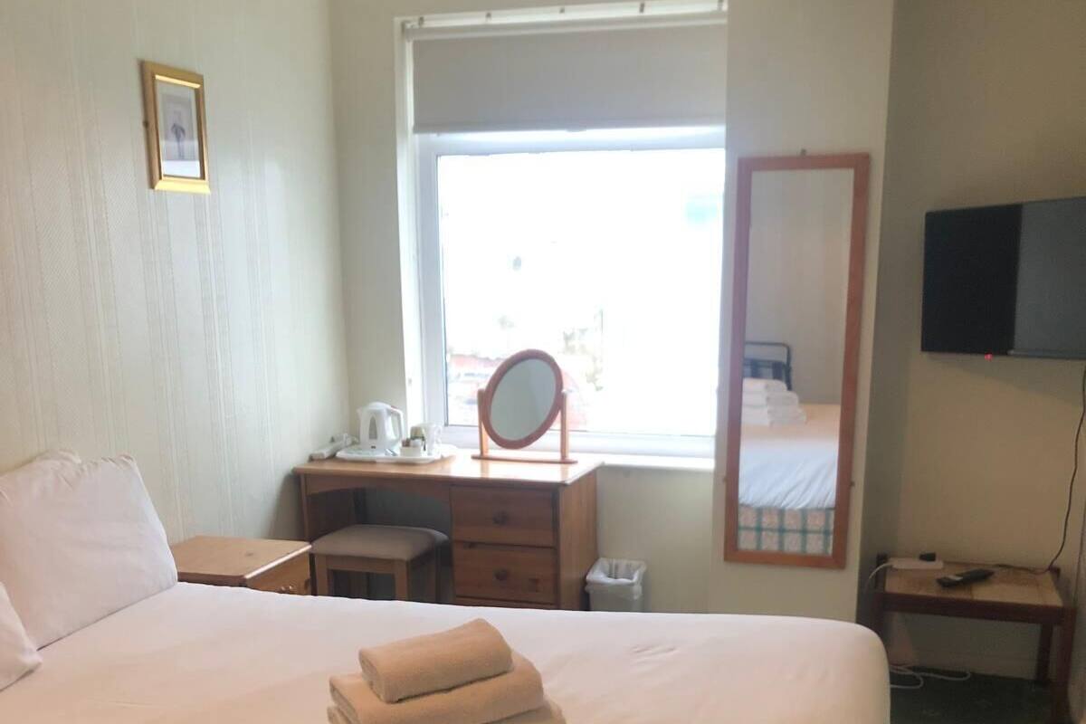 A neatly made bed with a pillow and folded towels, a bedside table with a lamp, a desk with a mirror, and a television on the wall.