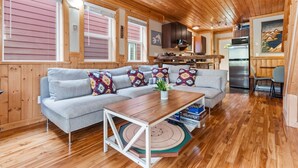 Ein gemütliches Blockhaushaus-Interieur mit grauem Sofa, Holzbalkendecke und einer Küchenzeile mit Kühlschrank und Schränken.