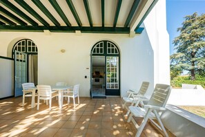 Un patio techado con mesa y sillas, una puerta que da a una cocina y vistas a un árbol y al paisaje.