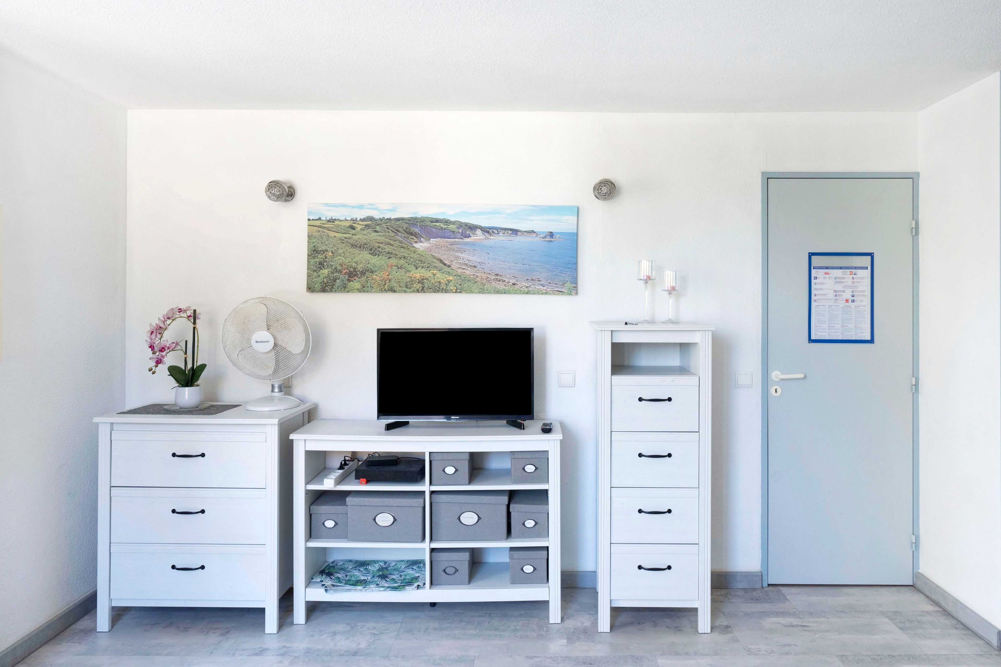 Una habitación con un escritorio blanco, un televisor de pantalla plana, una planta pequeña en maceta, un cuadro colgado en la pared y una puerta con un aviso.