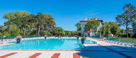 Una piscina con plataforma de baldosas, rodeada de árboles y un edificio con techo de tejas rojas.