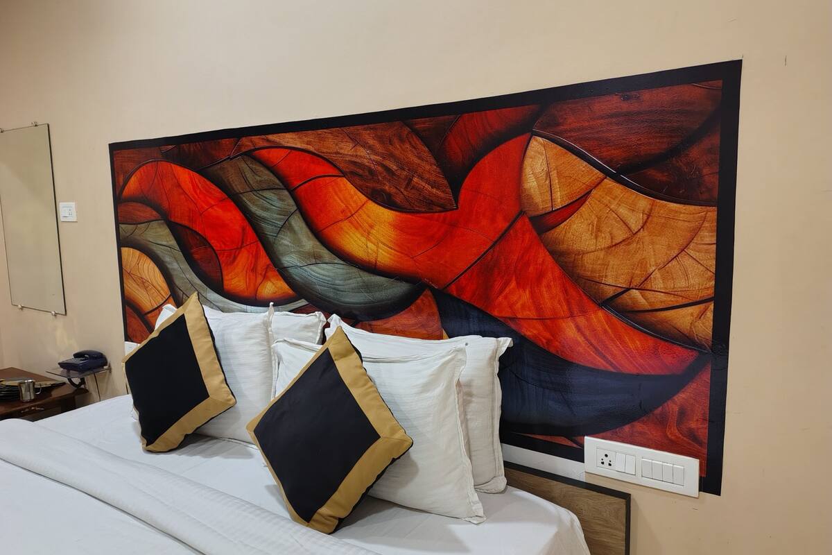 Una cama con ropa de cama blanca y dos almohadas negras y doradas, una mesita de noche con un teléfono y una lámpara, y un cuadro abstracto grande en la pared.