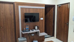 Une pièce avec un téléviseur fixé au mur, un mur en bois, une petite étagère avec des bouteilles d’eau et une bouilloire, et une porte avec une poignée.