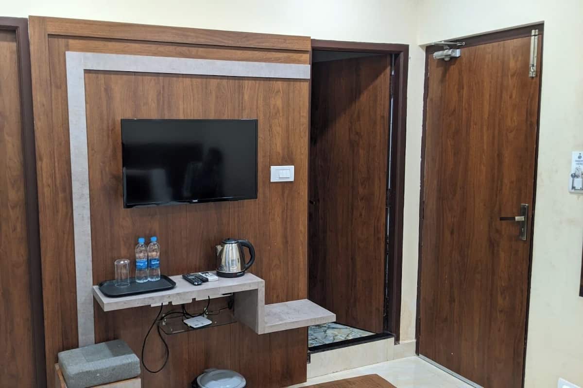 Una habitación con un televisor montado en una pared de madera, un pequeño estante con botellas de agua y una tetera, y una puerta con manija.