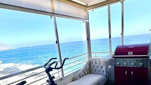 A balcony with a view of the ocean, a grill, a sofa, an exercise bike, and a small table with a plant. - South Beach Front Apartment (Antofagasta)