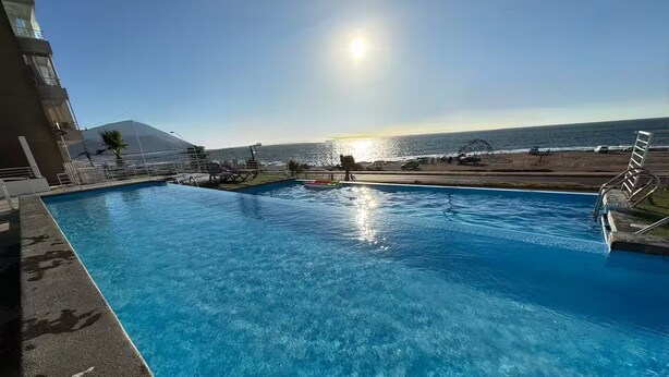 A swimming pool with clear blue water, a sunlit sky, and a view of the beach and ocean. - South Beach Front Apartment (Antofagasta)