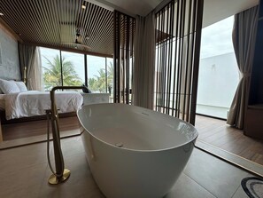 A modern bedroom with a large bathtub, a bed with white linens, and a view of palm trees through large windows. - Wyndham Hoi An Royal Beachfront Resort (Hoi An)