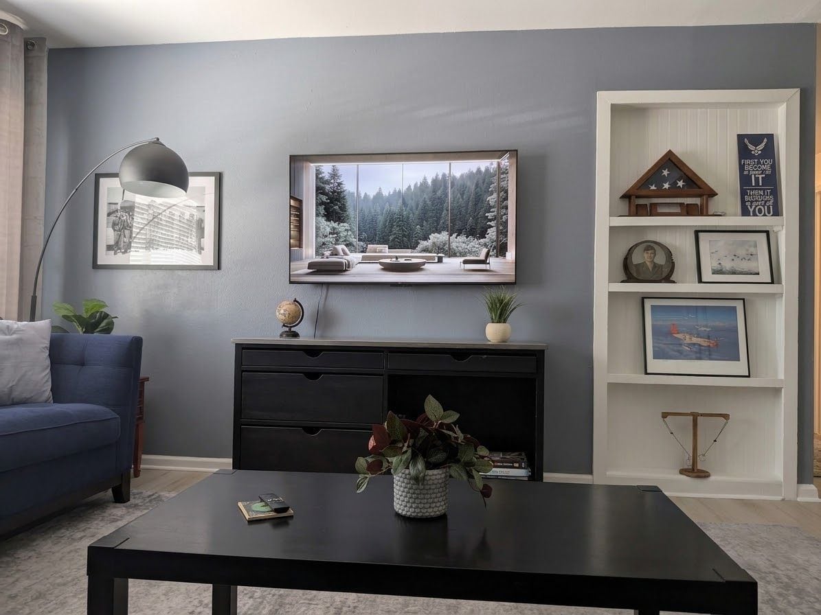 Een woonkamer met een bank, een bijzettafel met een plant, een aan de muur bevestigde televisie en een boekenkast met ingelijste foto’s en een klein huismodel.