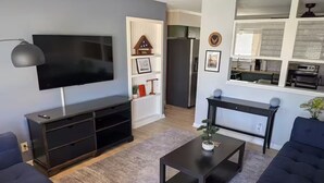 Una sala de estar moderna con un televisor de pantalla plana, sofás oscuros, una mesa de centro y una cocina visible a través de un umbral abierto.