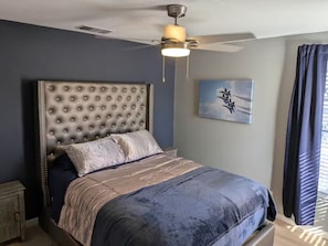 A bedroom with a tufted headboard, a ceiling fan, a bed with a striped bedspread, a nightstand, and a painting of an airplane on the wall. - Patriot's Post - just renovated near Lackland AFB (San Antonio)