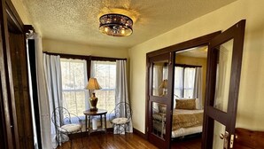 Une pièce avec un plancher en bois, une table avec une lampe, deux chaises, un lit recouvert d’une couvre-lit à motifs et une grande fenêtre avec des rideaux.