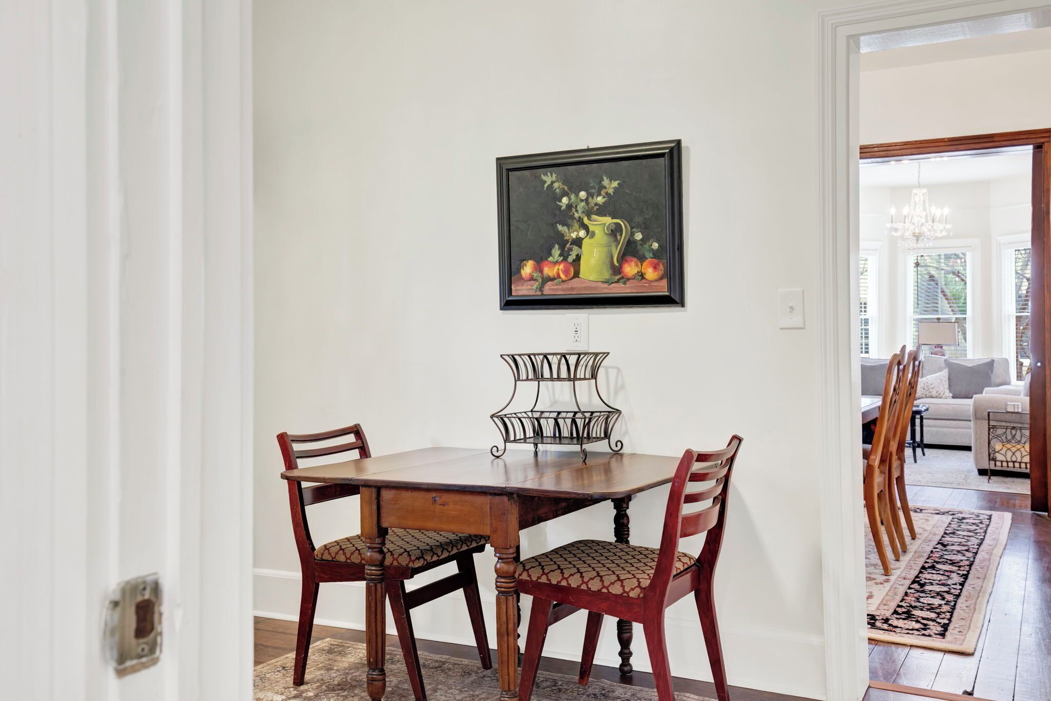 A dining area with a wooden table and chairs, a framed painting of fruit, and a decorative metal stand.