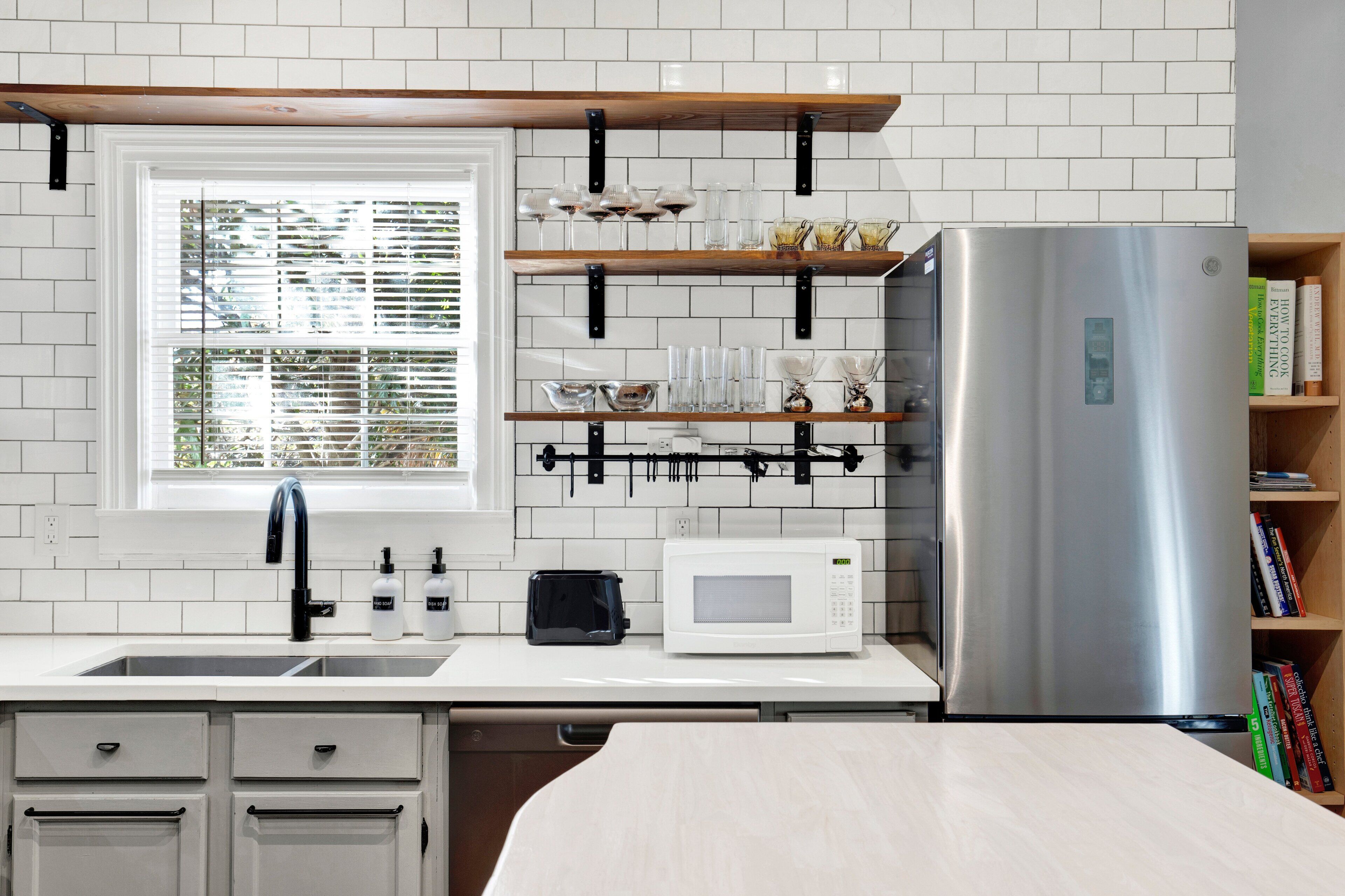 A modern kitchen with white cabinetry, a stainless steel refrigerator, a microwave, and a window with blinds.
