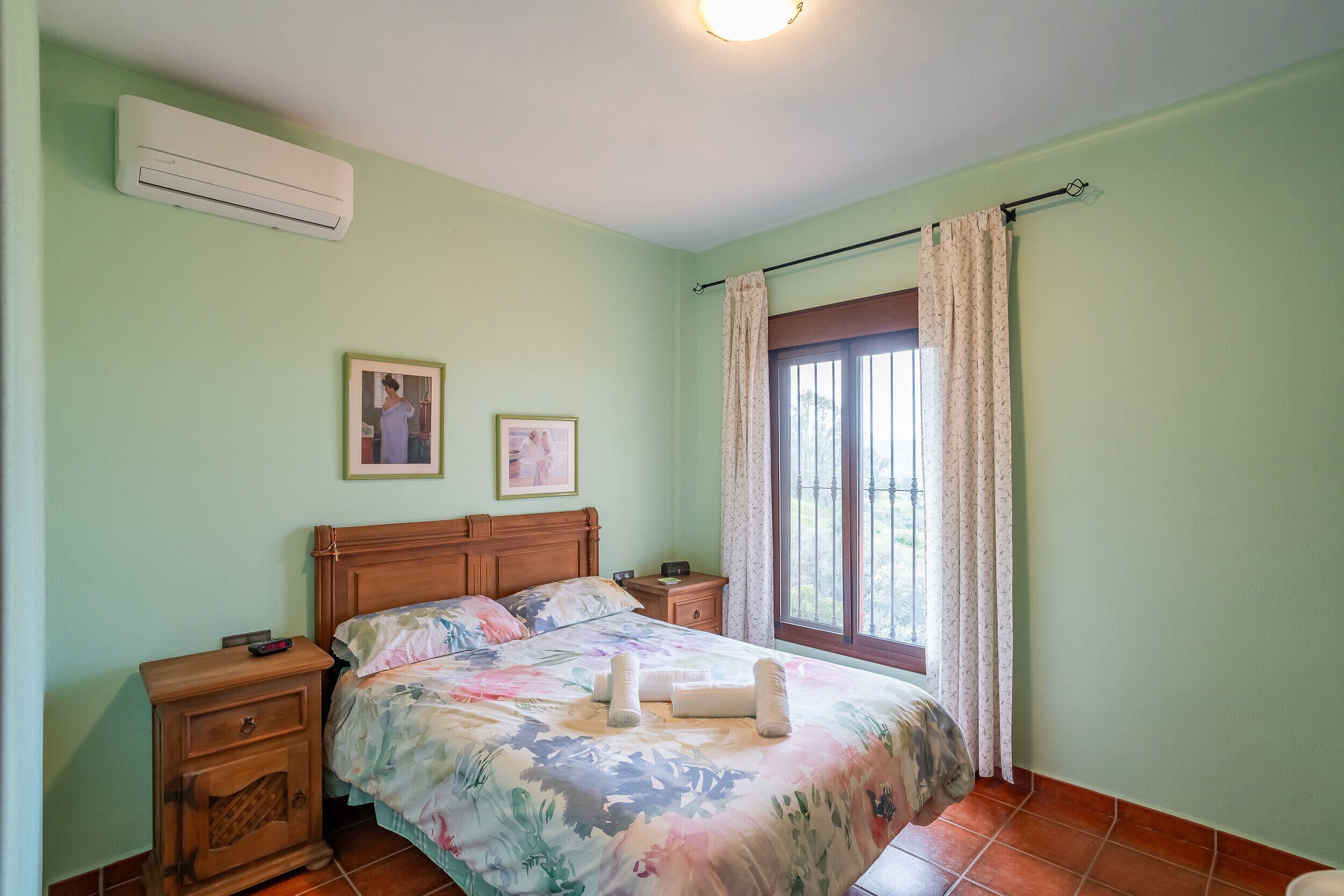 A bedroom with a bed, nightstands, an air conditioner, a window with curtains, and two framed pictures on the wall.