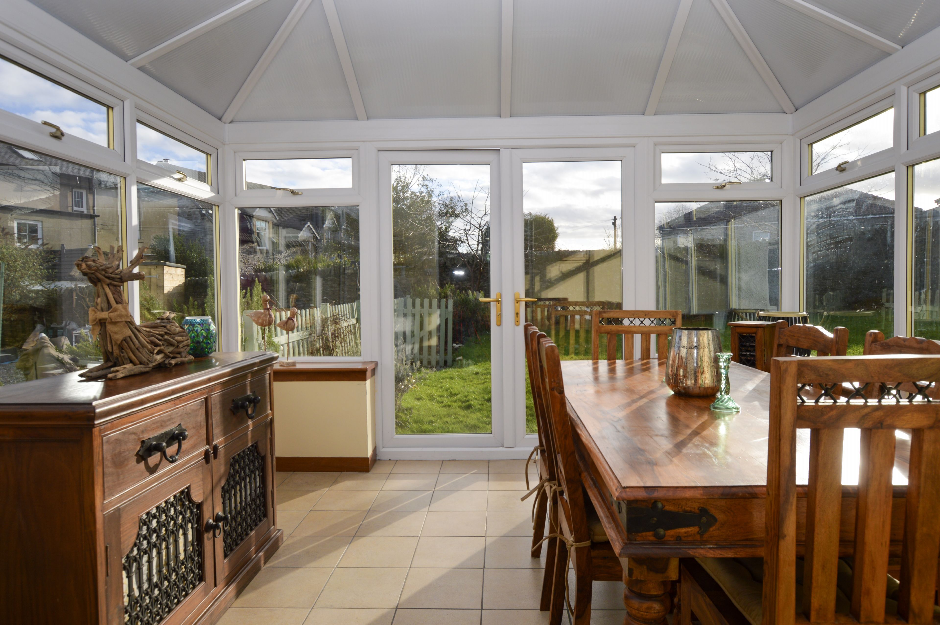 Ein Wintergarten mit einem Holzespeisetisch und -stühlen, einem Schrank mit Glastüren und Blick auf einen Garten durch große Fenster.