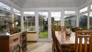 Un invernadero con una mesa y sillas de madera, un aparador con puertas de cristal y vistas a un jardín a través de grandes ventanales.