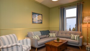 Una sala de estar con un sillón a rayas, un sofá gris con cojines estampados, una mesa de centro de madera y una ventana con cortinas.
