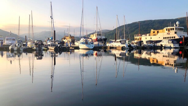 Un puerto deportivo con varias embarcaciones amarradas, agua tranquila que refleja los barcos y un paisaje montañoso al fondo.