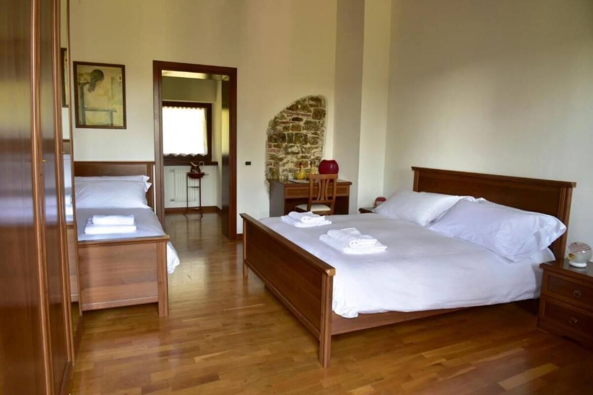 A bedroom with a wooden bed, a wooden nightstand, a desk with a chair, and a stone wall.