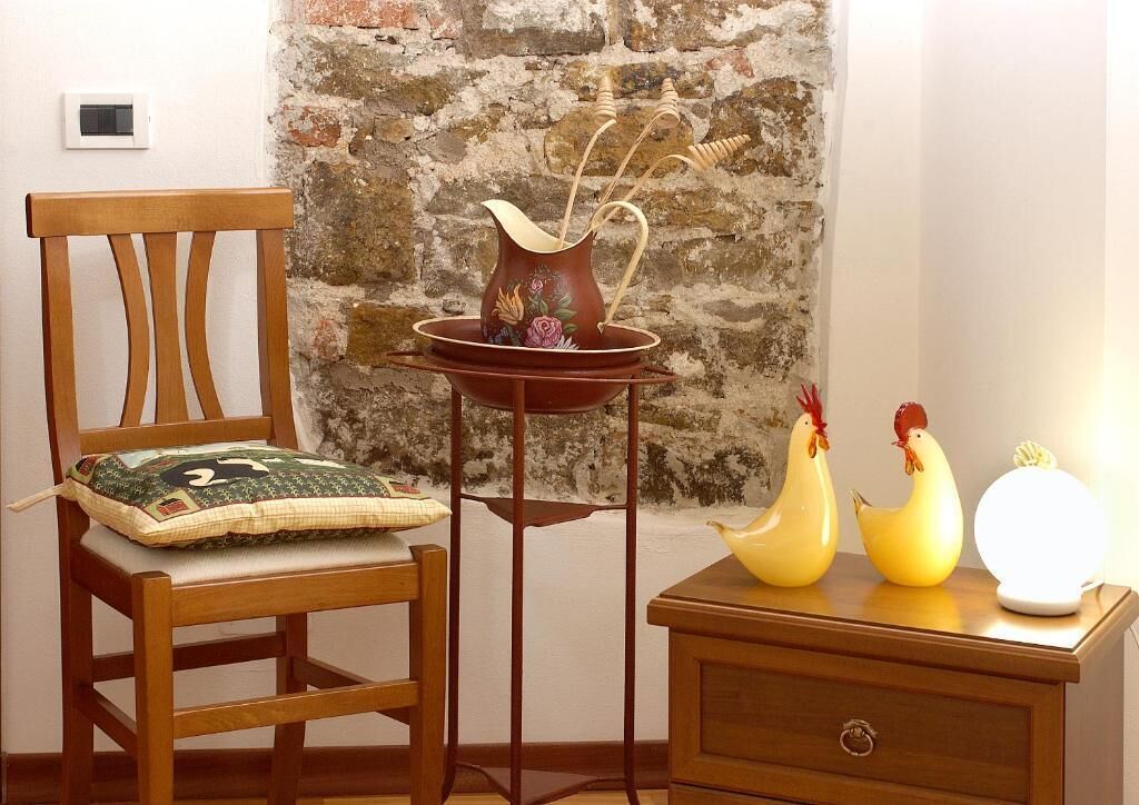 Una silla de madera con cojín, una mesita con un jarrón y adornos, y una mesita de noche con una lámpara y figuritas.