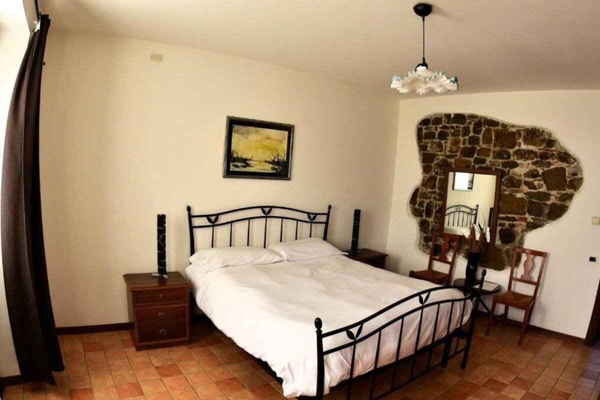 A bedroom with a bed, a nightstand, a chair, a small table, and a stone wall.