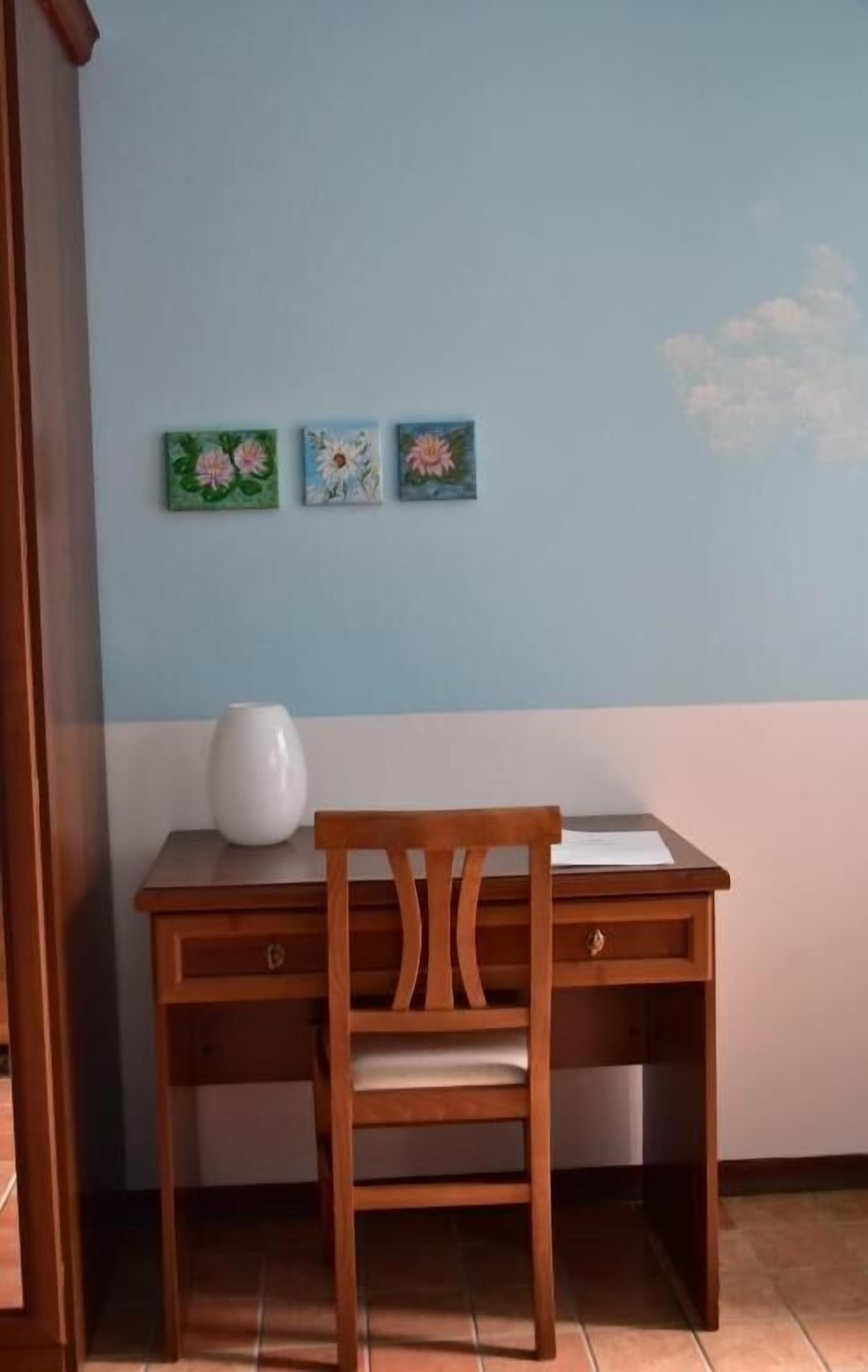 Un escritorio de madera con una silla, un jarrón blanco y tres cuadros con motivos florales en la pared.