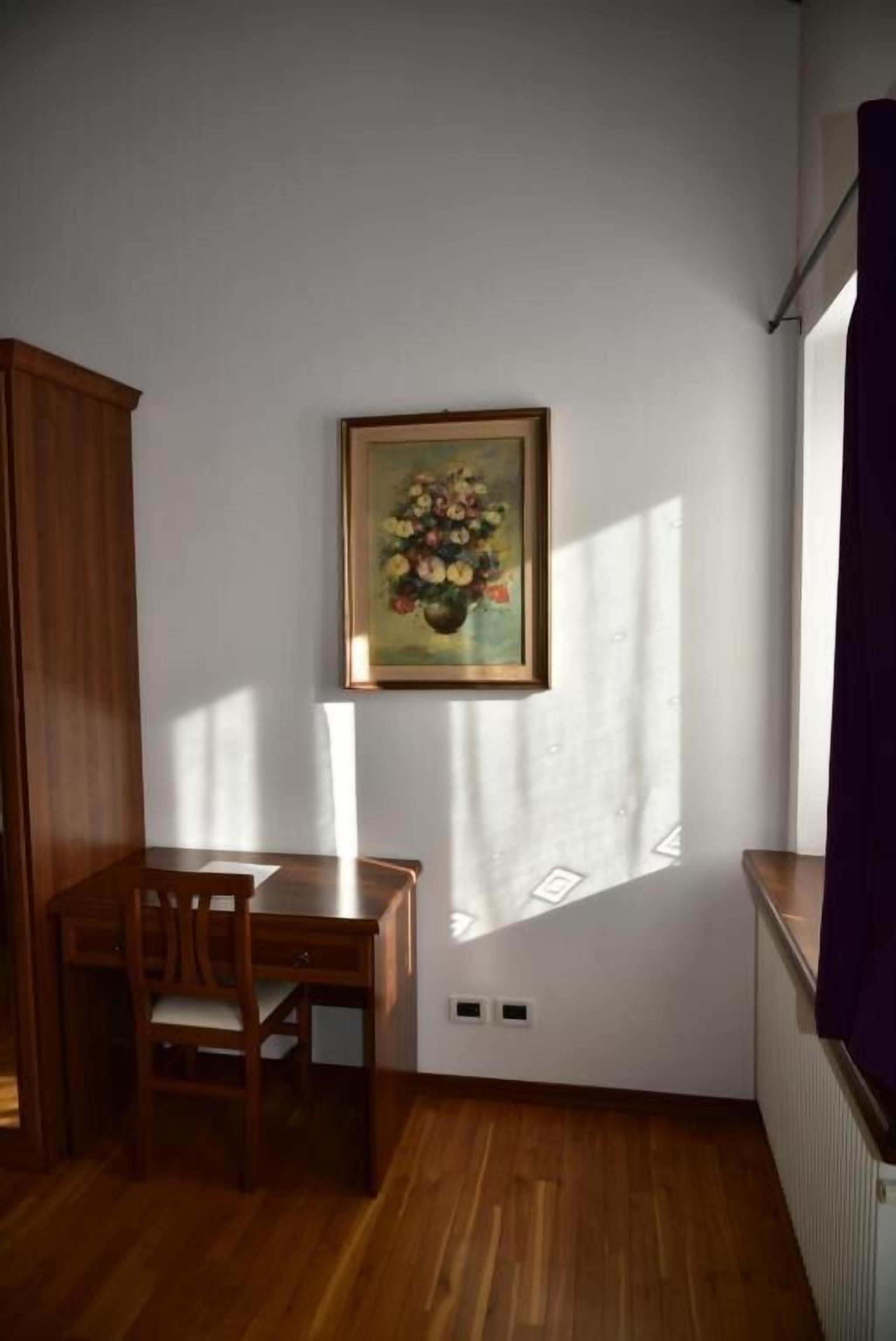 A room with a wooden table and chairs, a framed painting on the wall, and a window casting shadows on the floor.