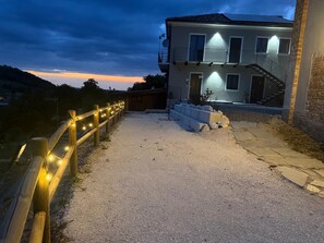 Un edificio con camino de grava, valla de madera con luces y una escalera que lleva a un balcón.