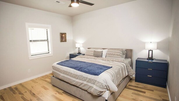 Une chambre à coucher comprenant un lit, deux tables de chevet, un ventilateur de plafond, une fenêtre et une petite table.