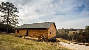 Une cabane en rondins avec un toit en pente, une terrasse en bois et un chemin de gravier menant à l’entrée.