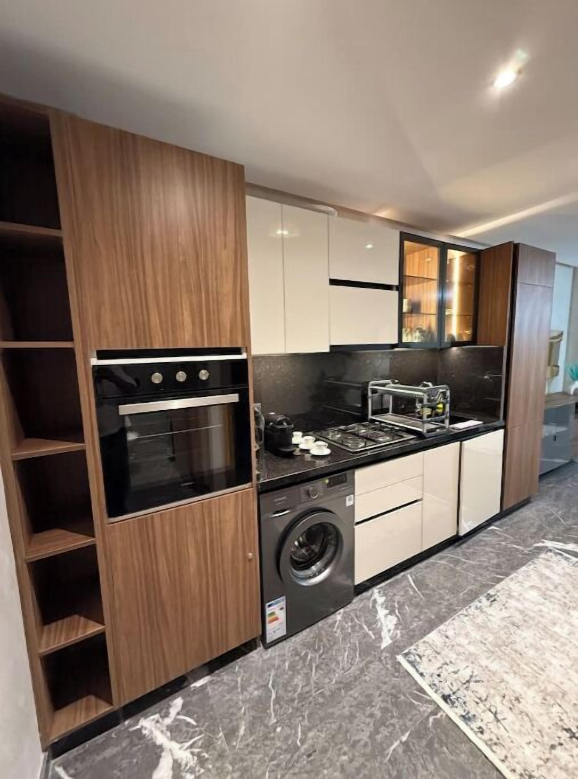 A modern kitchen with wooden cabinets, a built-in oven, a washing machine, and a marble countertop.