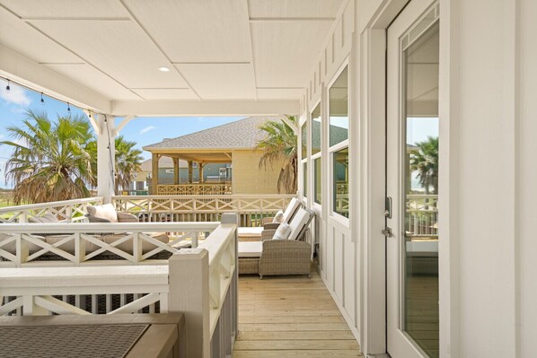 A covered outdoor area with a wooden deck, wicker furniture, and a view of a building with a sloped roof and palm trees. - Golf Cart, Elevator, Stylish, 12 guests - LaPerla (Bolivar Peninsula)