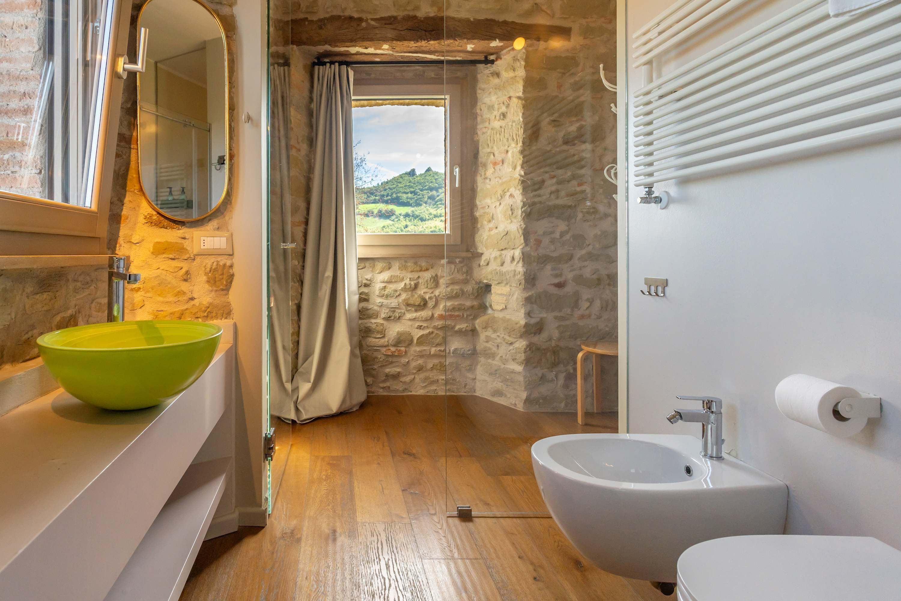 Un baño con pared de piedra, piso de madera, un espejo grande, un lavamanos verde, un bidé y vistas de un paisaje montañoso a través de una ventana.