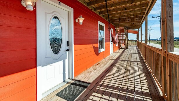 A covered porch with a wooden floor, a white door with a glass window, and a red exterior wall. - Cozy 2BR beach stay with a kitchenette—just a short walk to the sand. (Bolivar Peninsula)