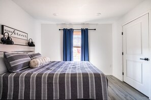 A bedroom with a bed, a bedside table, a lamp, a wall-mounted shelf with decorative items, and a window with curtains. - Cozy 2BR beach stay with a kitchenette—just a short walk to the sand. (Bolivar Peninsula)