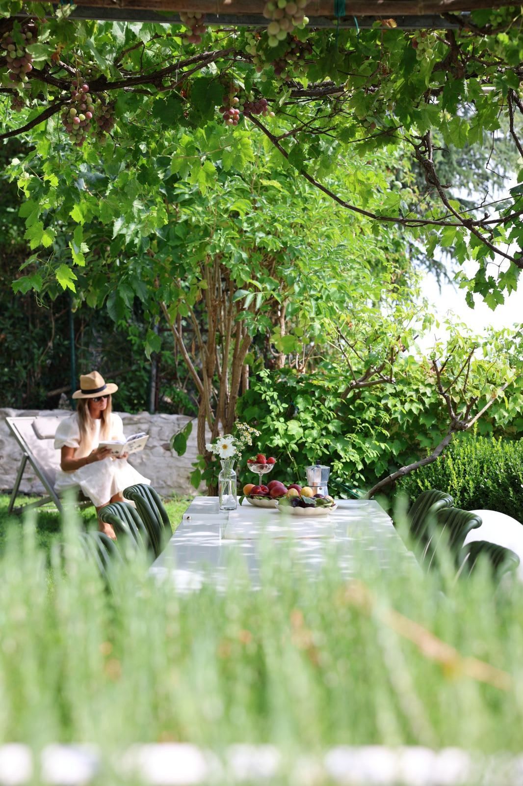 A person reading a book at a table with a garden view, a vase with flowers, and a fruit bowl.