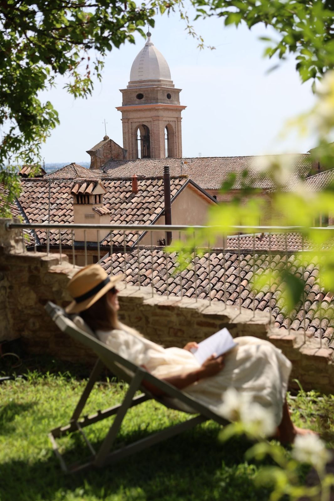 A person relaxing in a chair with a book, overlooking a historic building with a bell tower and tiled roofs.