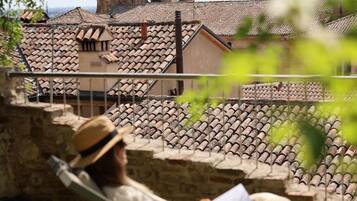 A person relaxing in a chair with a book, overlooking a historic building with a bell tower and tiled roofs.