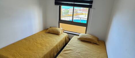 Une petite chambre avec deux lits simples, une fenêtre avec des stores et une vue sur une piscine.
