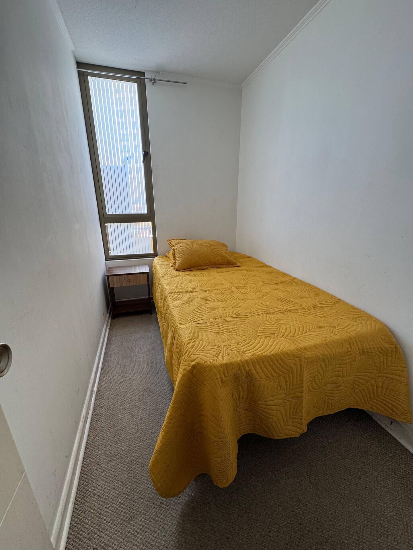 Une petite pièce étroite, avec un lit simple, une fenêtre avec des stores et une porte.