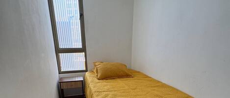 Une petite pièce étroite, avec un lit simple, une fenêtre avec des stores et une porte.