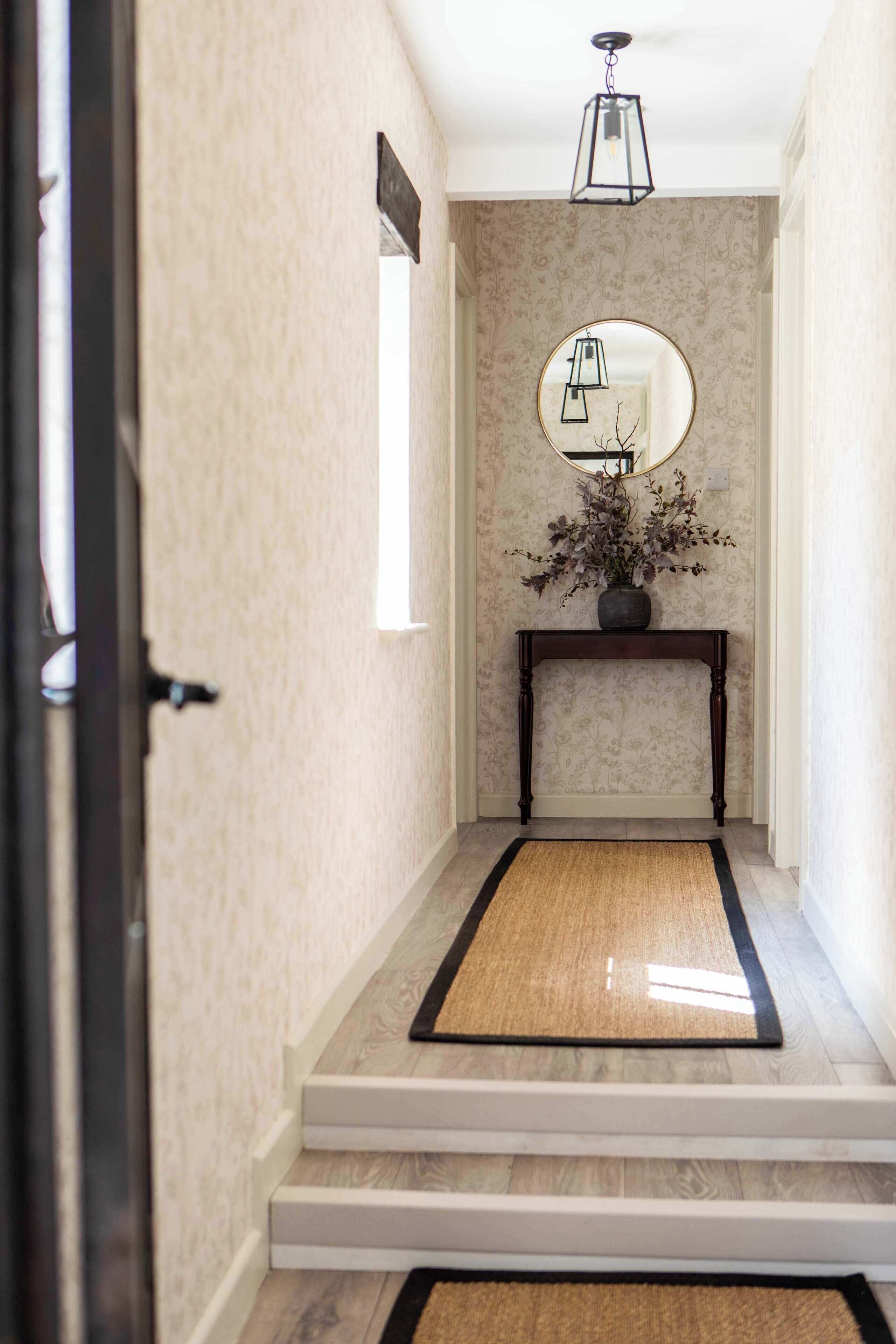 A hallway with a wooden floor, a black doormat, a wooden console table with a vase of flowers, a round mirror, and a hanging light fixture.