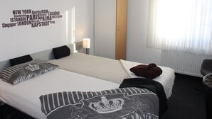 Habitación de hotel con dos camas, una pared con nombres de ciudades, una lámpara y una ventana con cortinas.