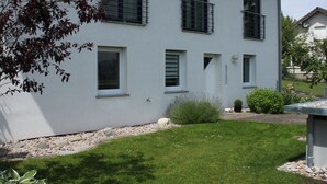 A modern two-story building with a white exterior, multiple windows, and a small garden area with a lawn and some shrubs.