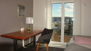 A hotel room with a bed, a desk, a chair, a window with a city view, and a painting on the wall.