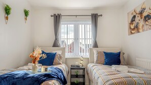 Un dormitorio con dos camas, una ventana con cortinas, una mesita con una planta y un cuadro en la pared.