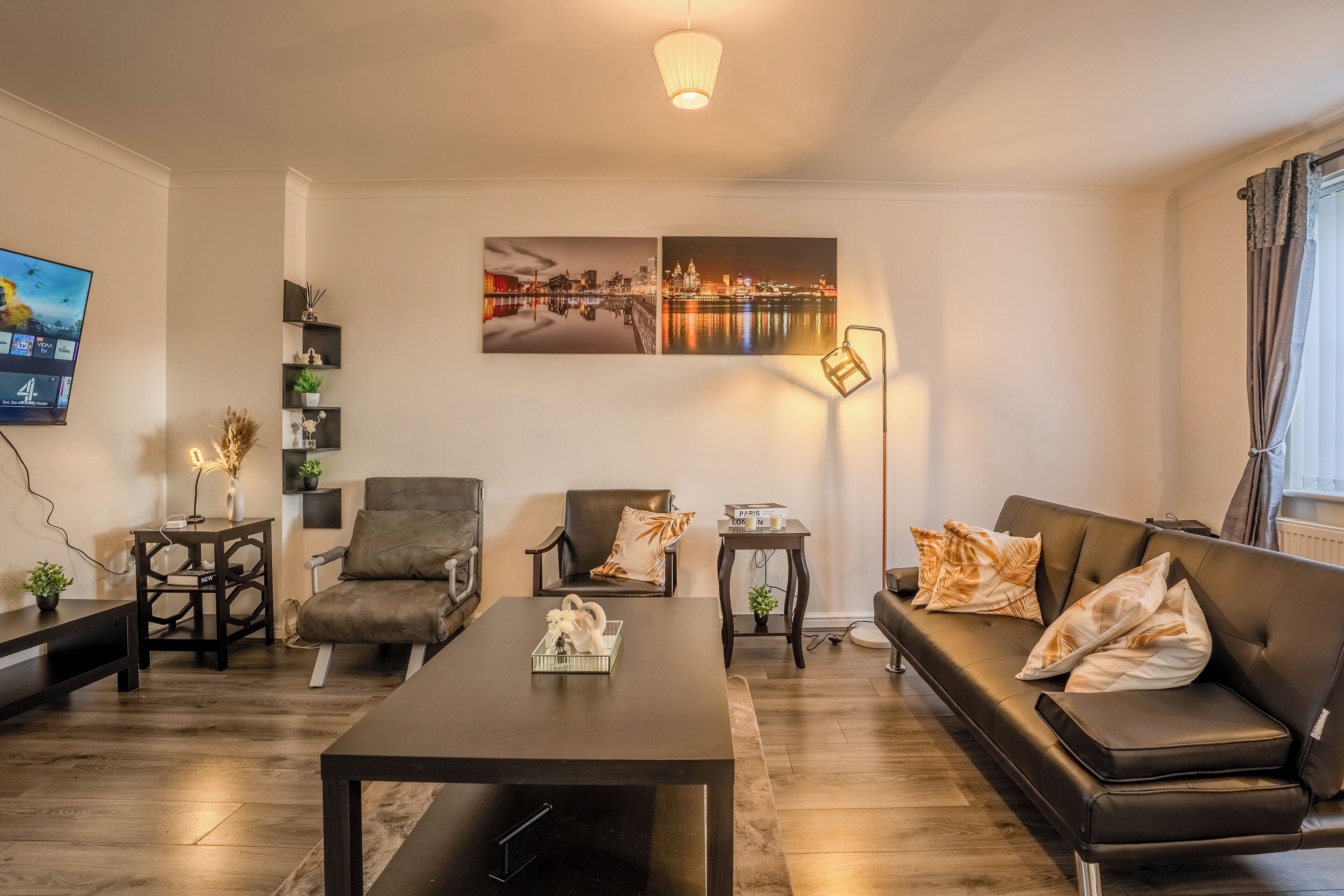 Un salon moderne avec un canapé, des fauteuils, une table basse et un téléviseur. Des objets décoratifs sont disposés sur des étagères et une lampe se trouve sur une table d’appoint.