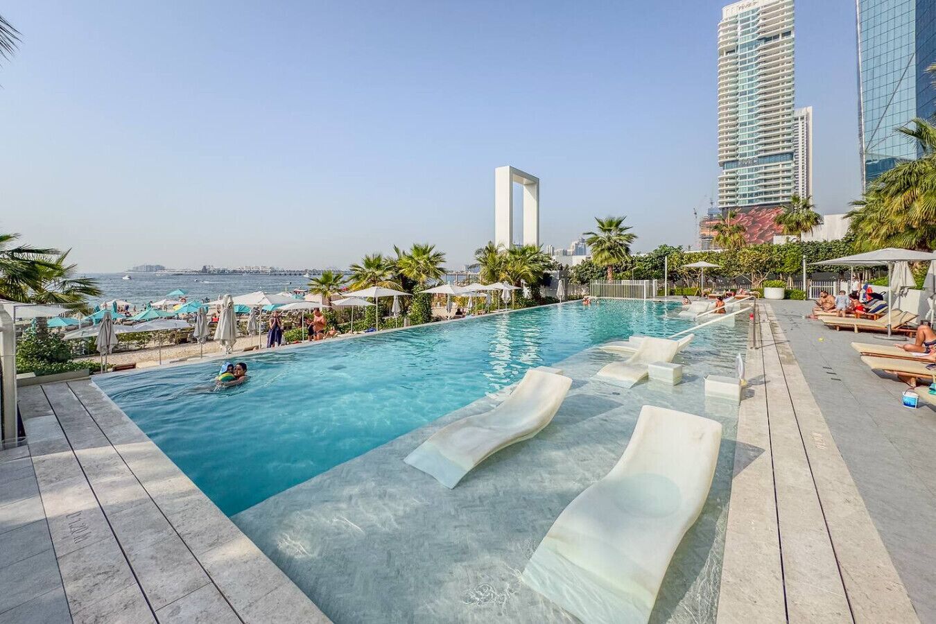 Ein Infinity-Pool auf dem Dach mit Liegestühlen, Palmen und einer Stadtkulisse im Hintergrund.
