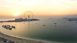 Un paysage urbain côtier avec une grande roue bien visible, une plage où se trouvent des gens, et des bateaux sur l’eau.